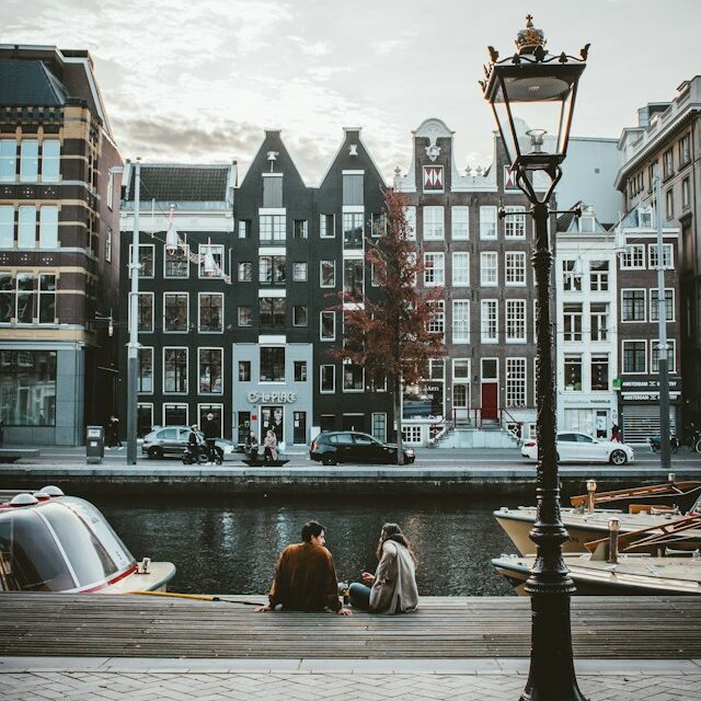 Two people talking while sitting on the edge of a canal with a row of buildings on the other side of the canal